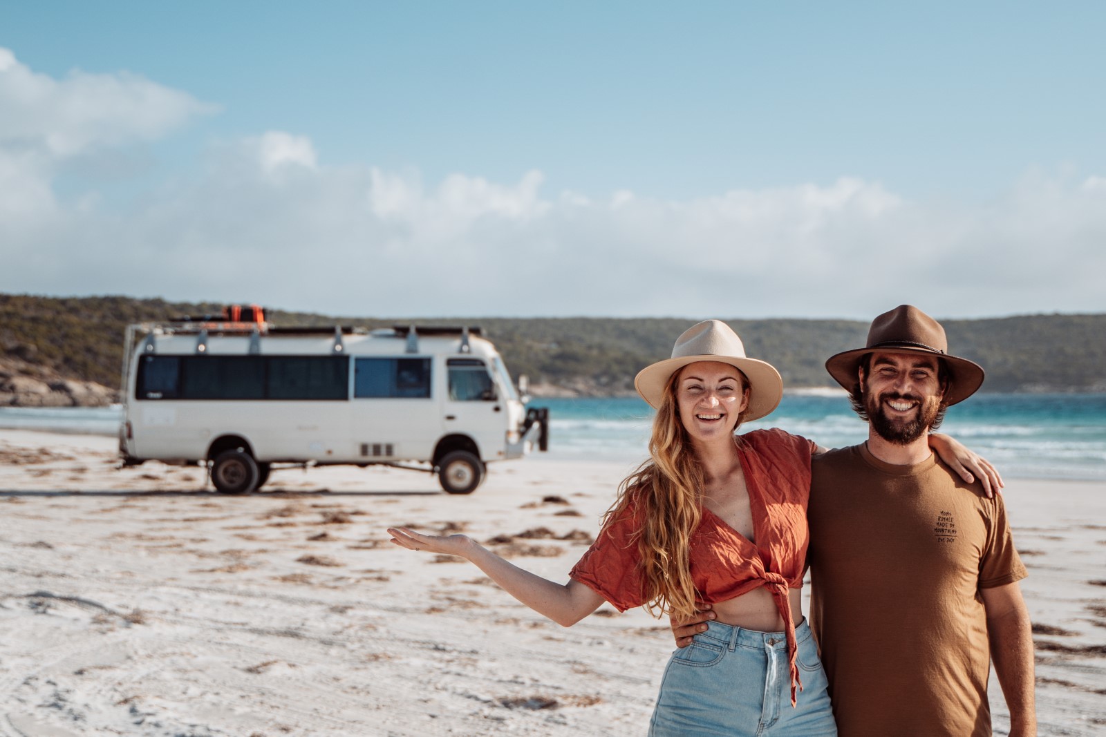 Chris and Beck of Salt and Charcoal stands on a sandy beach in Western Australia with Beck extending her arm showing the Salt and Charcoal camper van behind them.