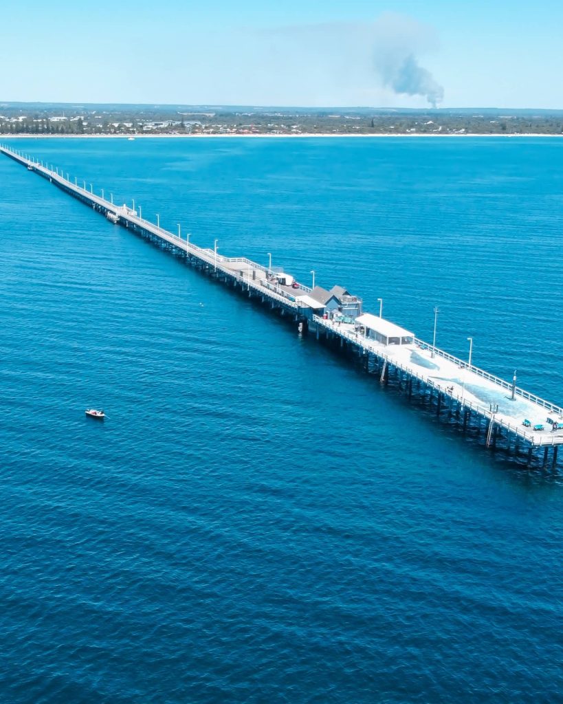 An aerial view of Busselton Jetty extending into the cobalt blue waters, with a single boat floating nearby.