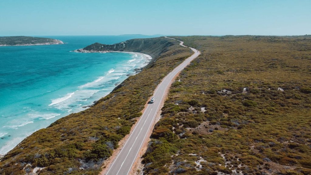 An aerial view of Esperance Coastal Drive in Western Australia, featuring the Salt and Charcoal van on a winding road with turquoise ocean on one side and dense shrubland on the other, under a clear blue sky.