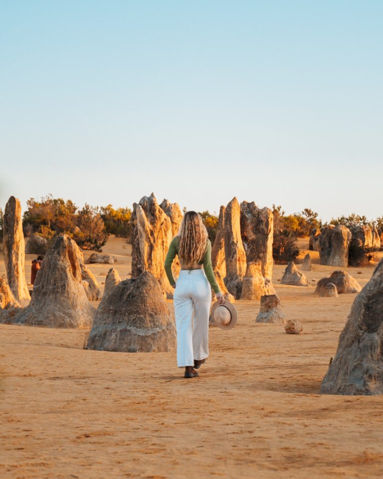 A woman walks among the limestone formations of the Pinnacles Desert in Australia, her back to the camera, holding a hat. The warm glow of the setting sun bathes the scene in golden light, casting long shadows on the sandy ground.