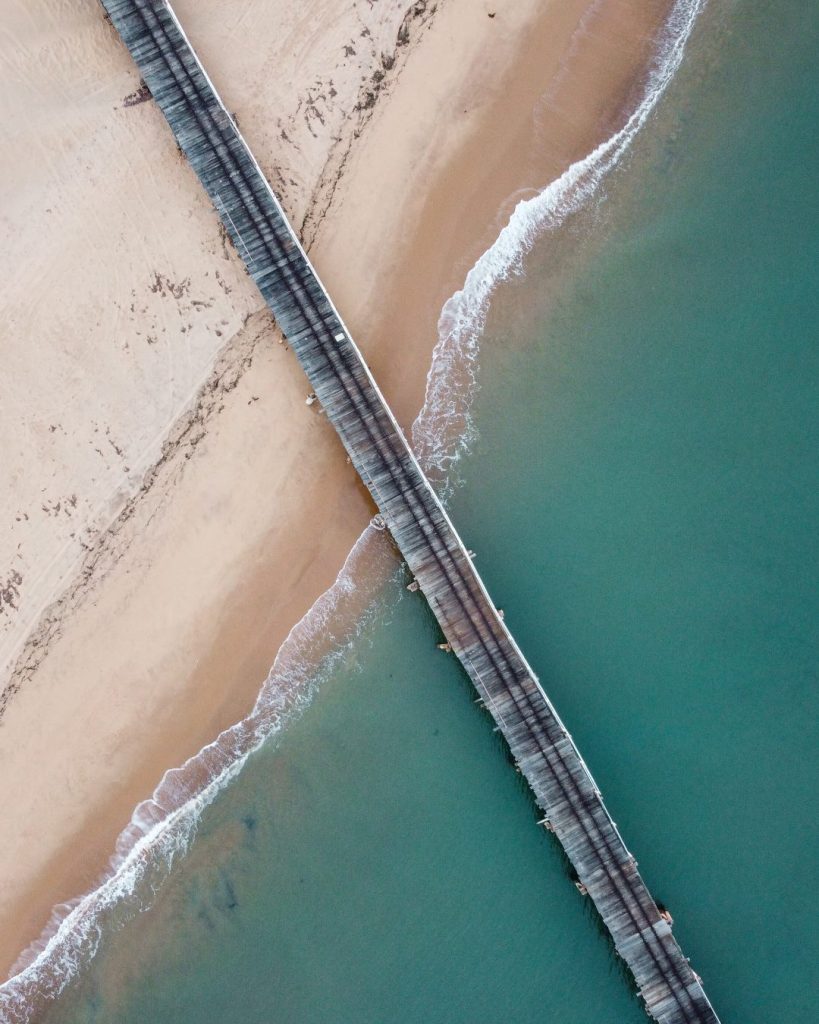 An aerial view of the Wharf in Carnarvon, Western Australia, shows the jetty extending straight across the image, dividing the sandy beach from the clear turquoise waters.
