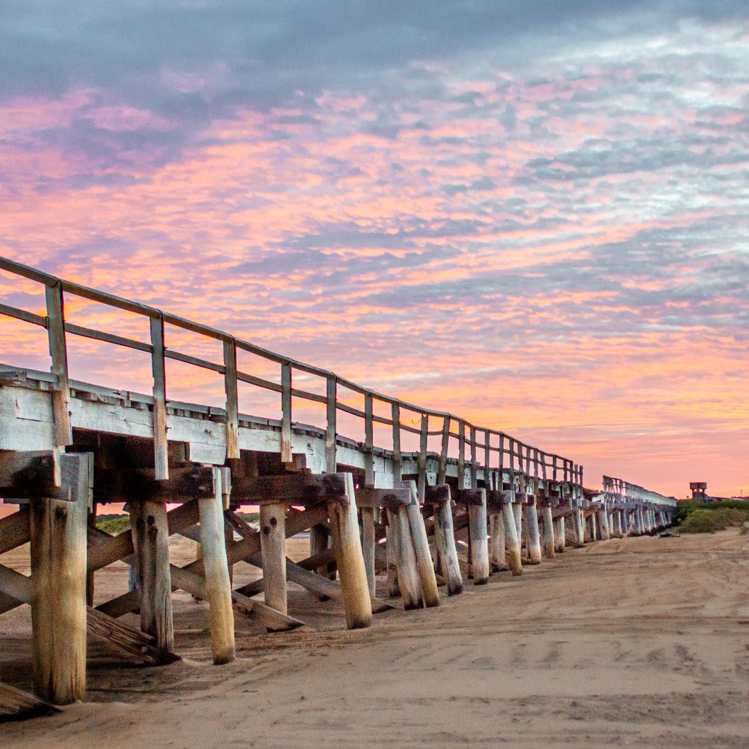 The historic Wharf in Carnarvon, Western Australia, stretches into the distance under a breathtaking sky painted with shades of pink and orange at sunset. The wooden structure of the jetty is prominently silhouetted against the vibrant colors, reflecting the calmness of the evening on this scenic coastline.