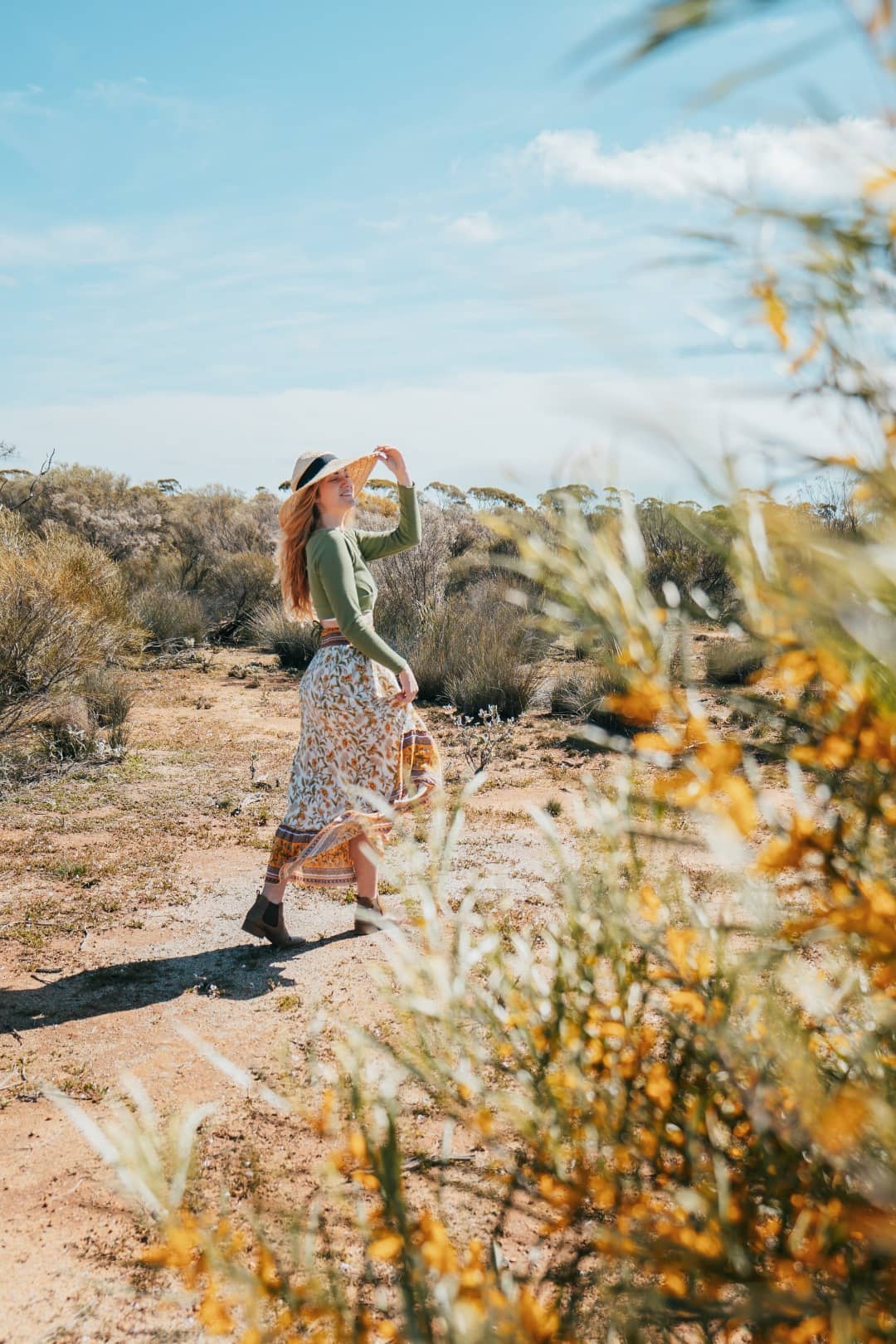 A woman in a long floral skirt and green top, holding onto her wide-brimmed hat as she wanders through a wildflower-strewn landscape in Western Australia. Golden wildflowers blur in the foreground, capturing the essence of a breezy day under the clear blue sky.