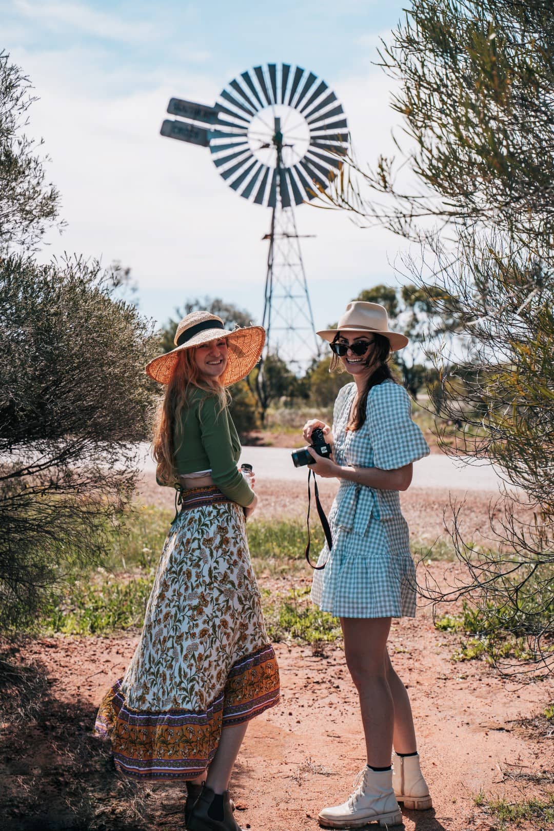 Two women in stylish sunhats are enjoying a sunny day in Western Australia’s countryside, with one holding camera. A traditional windmill can be seen in the background. The natural beauty of the bushes around them is indicative of a wildflower watch adventure.