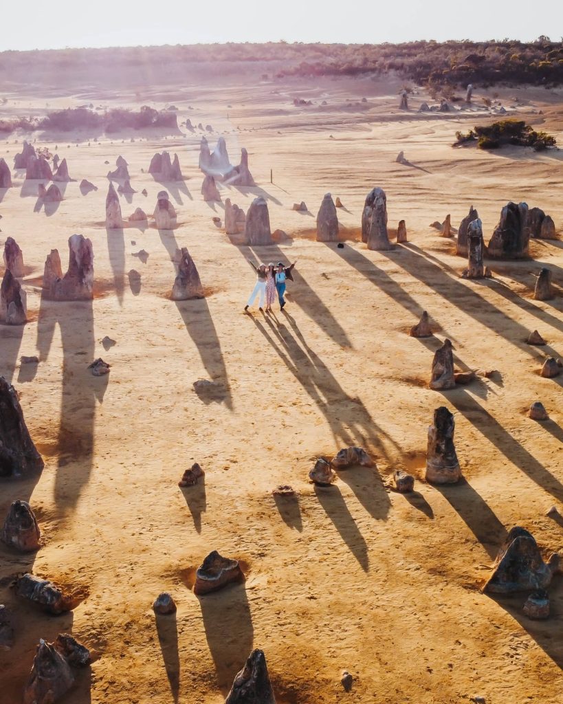 An aerial shot captures three happy people in the midst of the Pinnacles Desert, with the long shadows of the limestone formations stretching across the sandy terrain in the warm light of the setting sun.