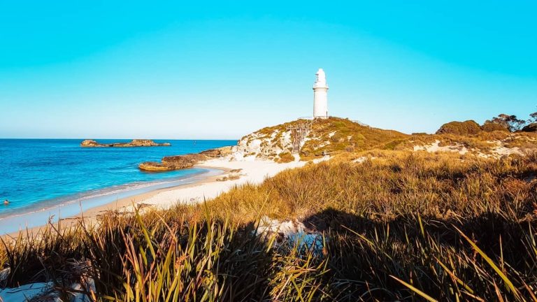 View of Bathurst Lighthouse on Rottnest Island, perched on a hill with sandy beaches and coastal vegetation in the foreground, under a clear blue sky and bright sunlight. One of the best locations for what to do on Rottnest Island