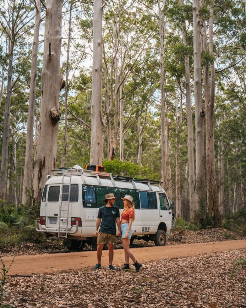 A couple stands next to a converted camper van parked on a dirt road, surrounded by tall trees in a lush forest of Boranup Drive in Margaret River, Western Australia.