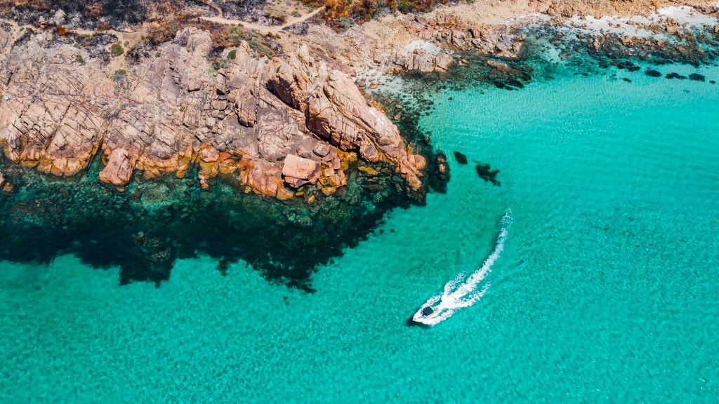 Aerial view of Castle Rock near Dunsborough, Western Australia, showcasing rugged reddish-brown cliffs meeting clear turquoise waters with a small boat cruising along the serene coastline. Perfect for those exploring things to do in Dunsborough.