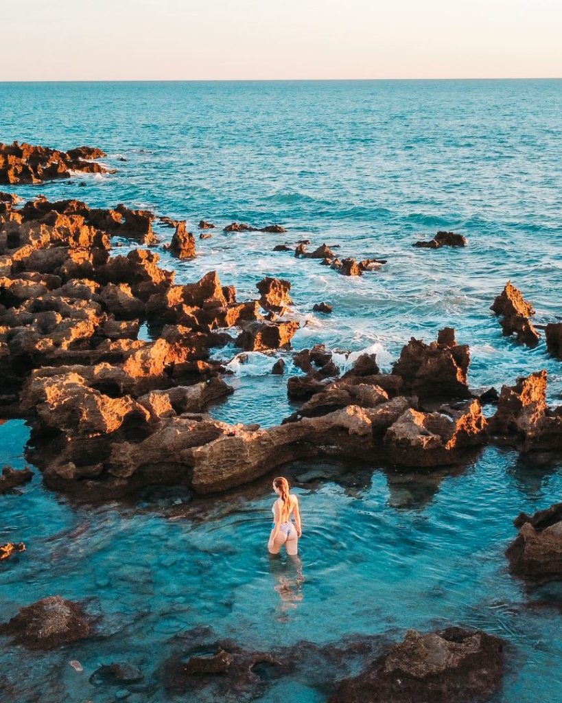 During sunset, a woman wades through the clear, shallow waters of the rock pools at Coconut Well in Broome, Western Australia.