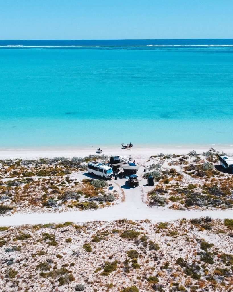 Remote beachfront campsite at Coral Bay with campervans and a 4WD parked beside white sand and clear turquoise water during a road trip from Perth to Exmouth. A small group stands near the shoreline with boats pulled up on the beach highlighting an off the beaten path stop in Western Australia.