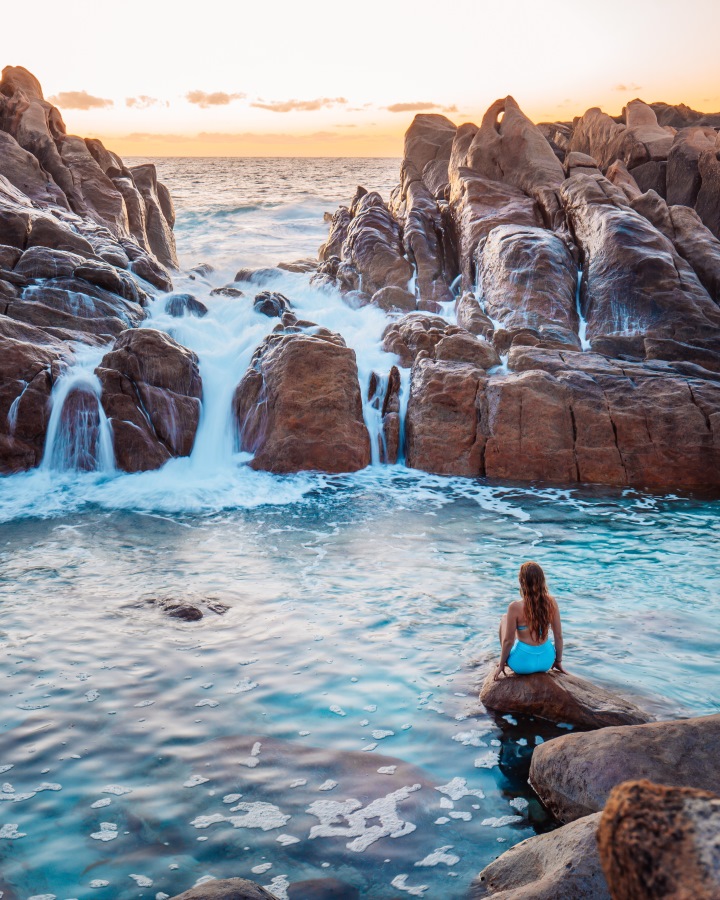 A woman in a blue bikini sits on a rock at Injidup Natural Spa in Western Australia, gazing at the cascading waves flowing over the rocky formations into the turquoise pool. This scene shows things to do in Dunsborough