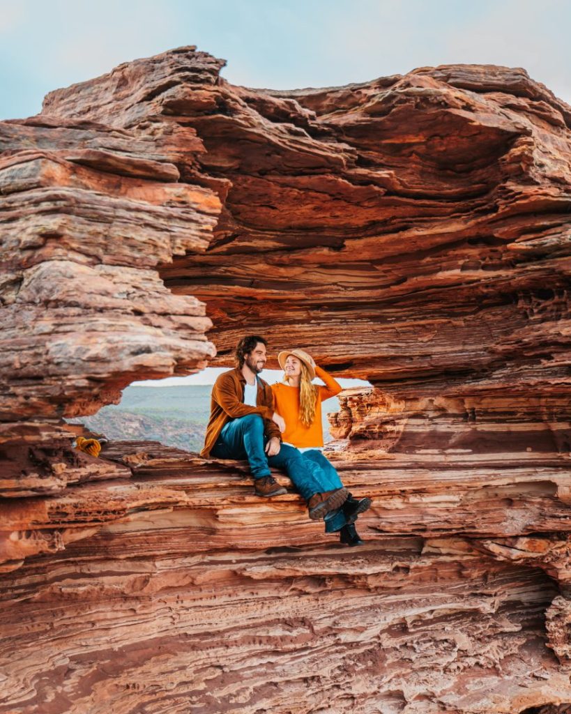 A couple sits within Nature's Window, a natural rock formation in Kalbarri National Park, Western Australia. They are framed by the layered red sandstone, enjoying the scenic views beyond.