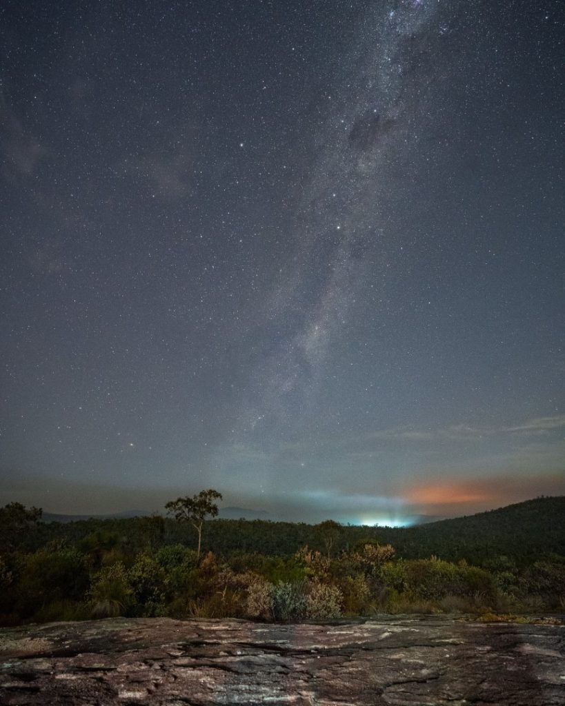 The image shows a breathtaking night sky filled with stars and the Milky Way stretches above a rugged landscape of trees and hills with a distant glow of city lights in Perth, Western Australia.