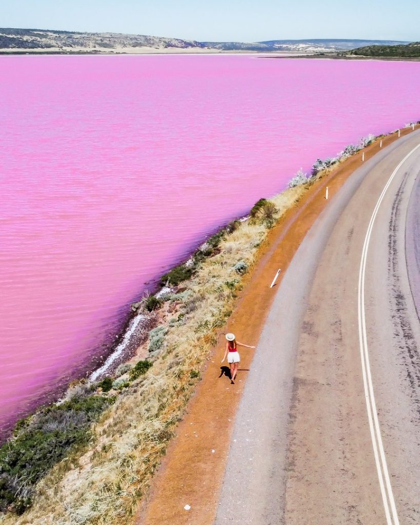 A woman in a red top and white skirt walks along a road next to the strikingly pink waters of Hutt Lagoon in Kalbarri, Western Australia.