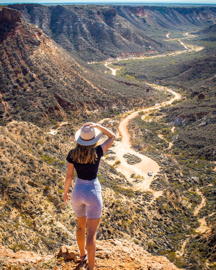 A woman stands at the edge of a cliff in Shothole Canyon, Cape Range National Park, looking out over a winding dirt road that carves through the rugged, arid canyon landscape. She wears a white hat and casual clothing, her hand holding the hat against the bright, sunny backdrop.