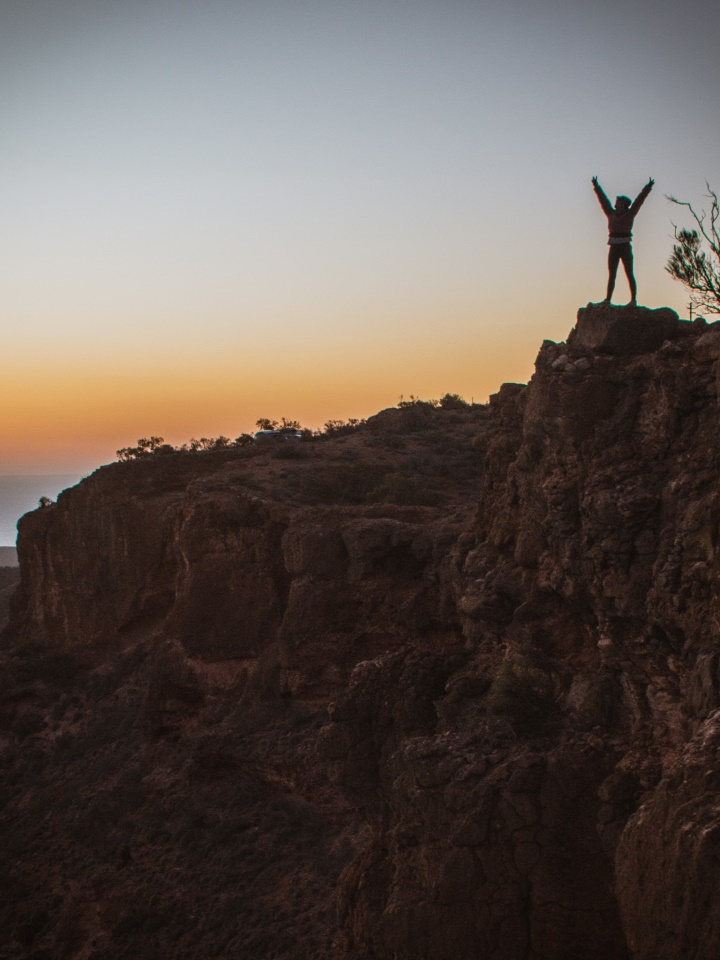 At dusk, a silhouette of a person with arms raised triumphantly stands atop a rugged cliff in Charles Knife Canyon, Cape Range National Park, Western Australia. The background features a soft gradient of sunset colors transitioning from warm orange to deep blue.