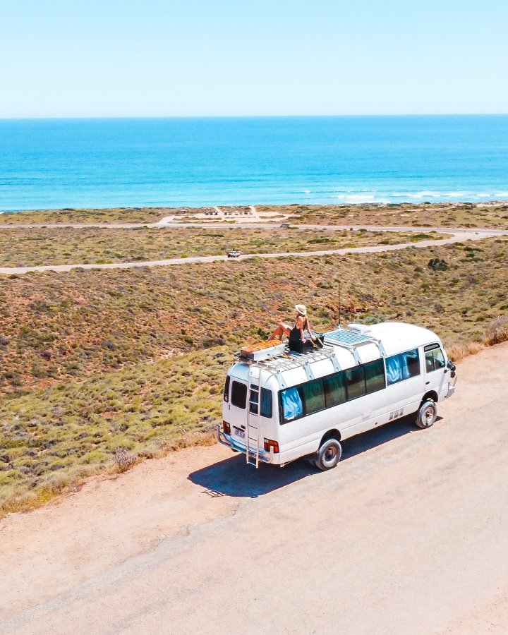 Aerial view of a woman sitting atop a white van parked on a coastal road in Exmouth, with a panoramic backdrop of Cape Range National Park rugged coastline and the blue ocean. The sunny day highlights the vivid colors of the landscape.