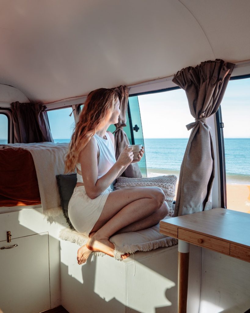 A woman sits inside a camper van, enjoying a cup of coffee while looking out at the ocean through the window. It shows a comfortable road trip in Western Australia.