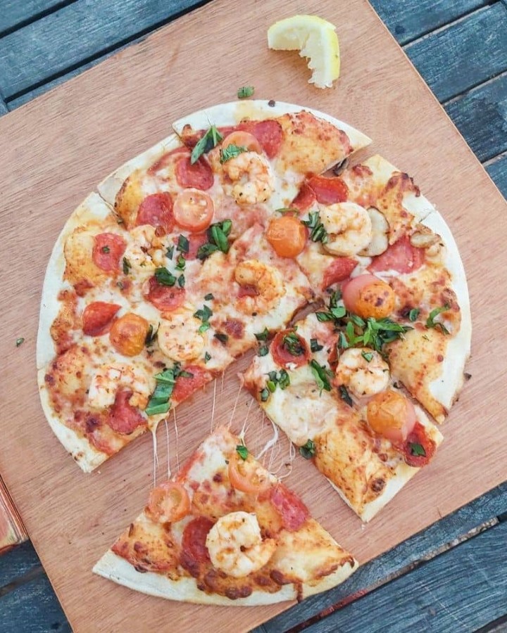 Overhead shot of a freshly baked seafood pizza on a wooden table, from the Whalebone Brewery in Exmouth, WA. The pizza features toppings of shrimp, sliced pepperoni, cherry tomatoes, and fresh basil. A slice of lemon sits on the side.