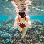 A snorkeler in a red swimsuit gives a thumbs-up while exploring the underwaters of Rottnest Island. The clear blue water reveals a thriving coral reef below. It hints snorkelling as one of the activities included in the best Rottnest Island tours