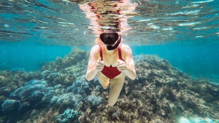 A snorkeler in a red swimsuit gives a thumbs-up while exploring the underwaters of Rottnest Island. The clear blue water reveals a thriving coral reef below. It hints snorkelling as one of the activities included in the best Rottnest Island tours