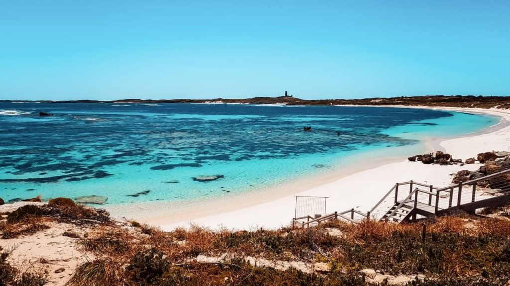 A stunning view of The Basin on Rottnest Island features crystal-clear turquoise waters and a white sandy beach. Wooden stairs lead down to the beach, showcasing what to do on Rottnest Island.