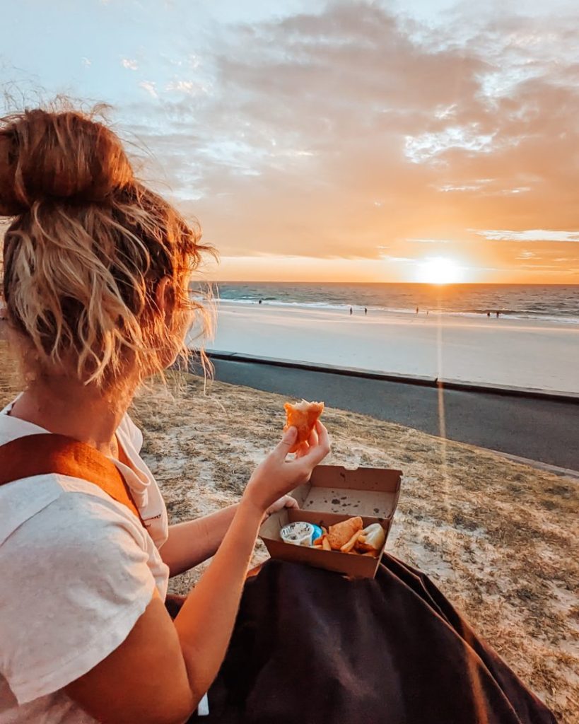 A woman enjoying fish and chips while watching the sunset at City Beach in Perth, Western Australia.