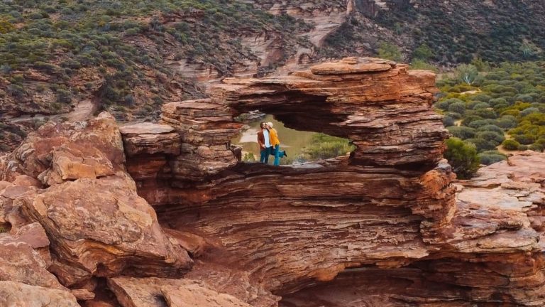 Two people stand at Nature's Window, a natural rock arch in Kalbarri National Park, Western Australia, with rugged landscape visible through the opening. It showcases of the things to do in Kalbarri, Western Australia.