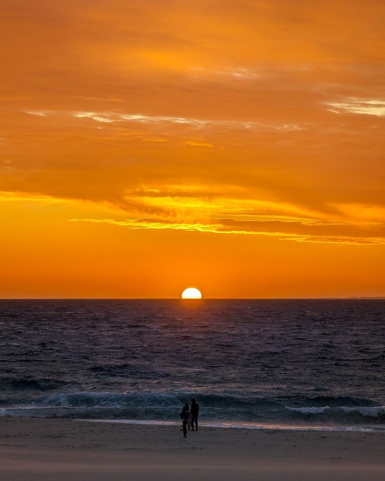 Sunset at City Beach in Perth, Western Australia, with the sun dipping below the horizon and two people walking on the beach.