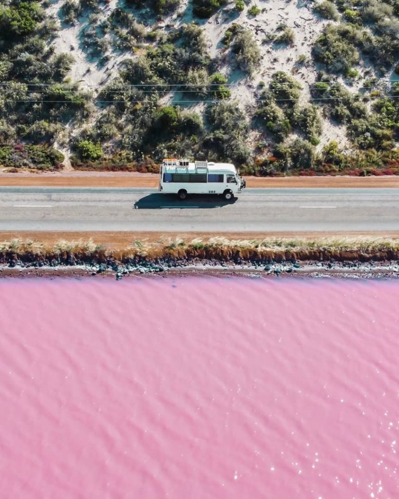 A camper van drives along a road beside the pink waters of Hutt Lagoon, also known as Pink Lake, in Kalbarri, Western Australia.