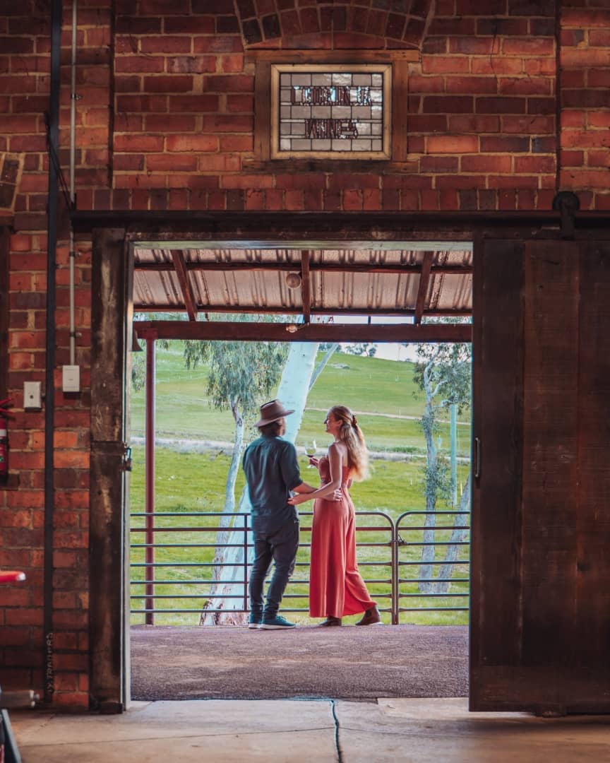 A couple in a wine-tasting experience in Swan Valley. They stand on a balcony, framed by an open barn door, looking out over a lush, green landscape. The woman wears a long, red dress while the man wears a hat and a dark shirt; they share a peaceful moment drinking wine together in Swan Valley, Perth, Western Australia.