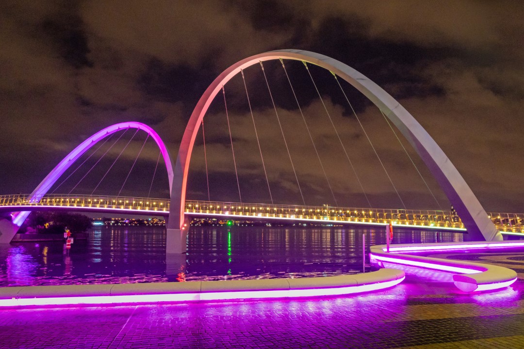 The Elizabeth Quay Bridge in Western Australia is illuminated at night with vibrant purple and yellow lights reflecting on the water below.