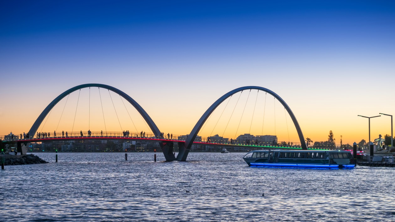 The Elizabeth Quay Bridge in Perth, Western Australia, at sunset with silhouettes of people walking on the bridge and a ferry with blue lights on the water below.