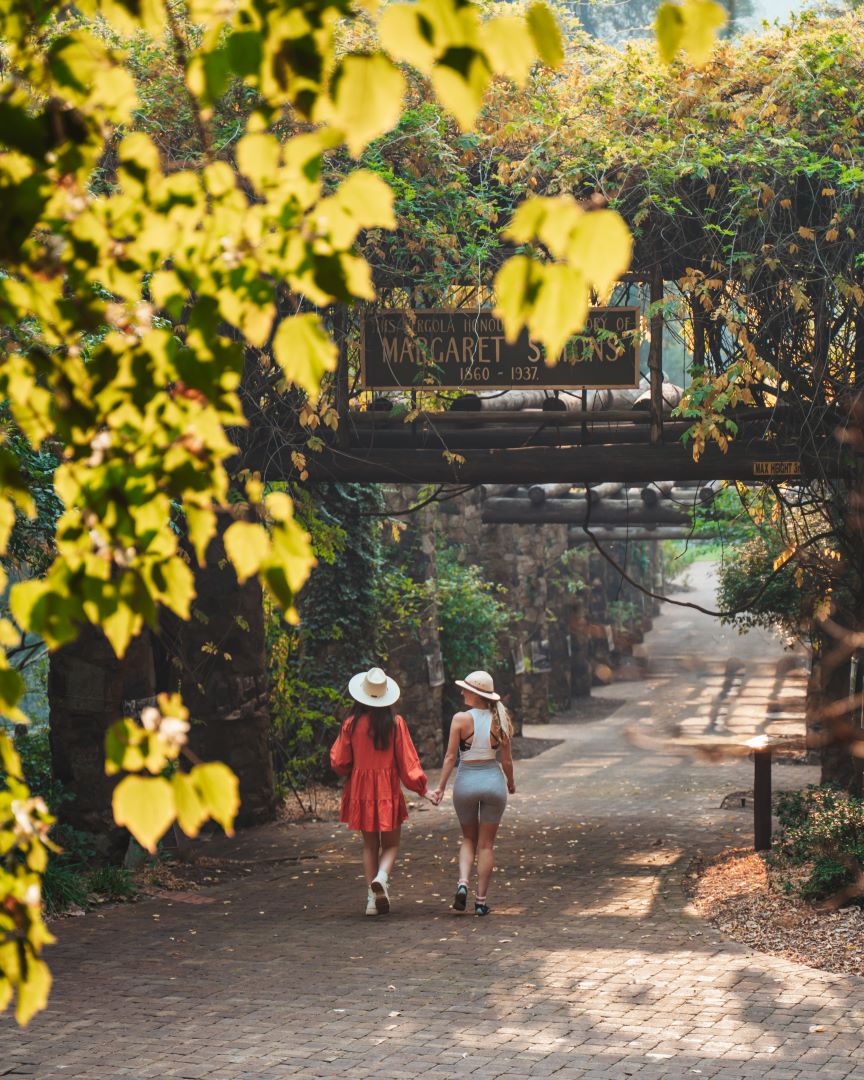 Two women in hats walk hand-in-hand down a tree-lined path towards a vine-covered archway of margaret simons pergola in Araluen Botanical Park Westen Australia.