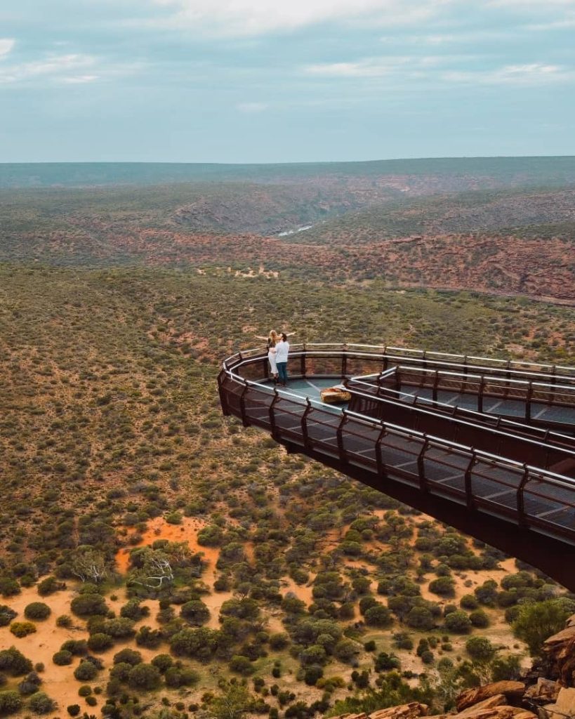 Drone shot of the Kalbarri Skywalk in Western Australia, with two people standing on the lookout platform, overlooking a vast, rugged landscape of canyons and bushland.