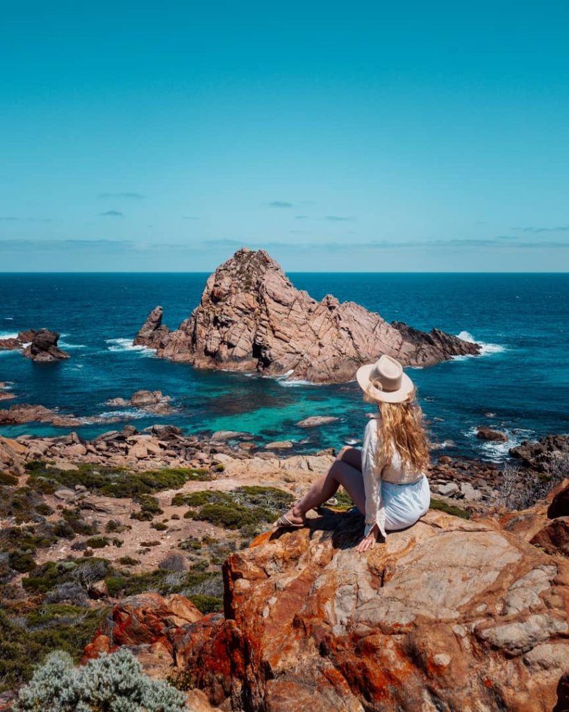 A woman in a hat sits on a rocky ledge overlooking Sugarloaf Rock, Western Australia. It hints at one of the things to experience on the best tours in Margaret River.