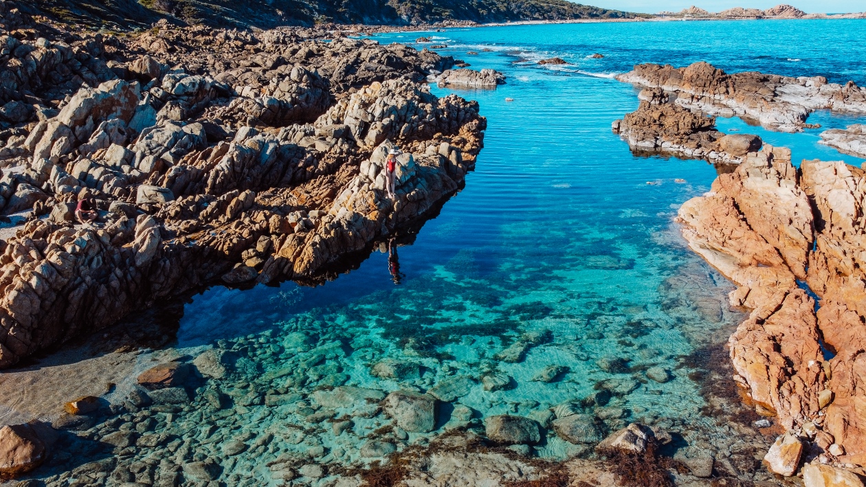 An aerial view of The Aquarium in Yallingup, Western Australia showing clear blue waters surrounded by rugged rocks with a woman standing on one near the water