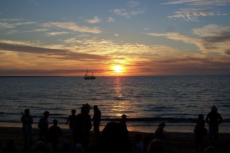 Sunset over the ocean at Mindil Beach in Darwin, Northern Territory, with silhouettes of people on the shore, as the final destination of a road trip from Adelaide to Darwin. A sailboat is visible on the water, framed by the vibrant colors of the setting sun and scattered clouds in the sky.