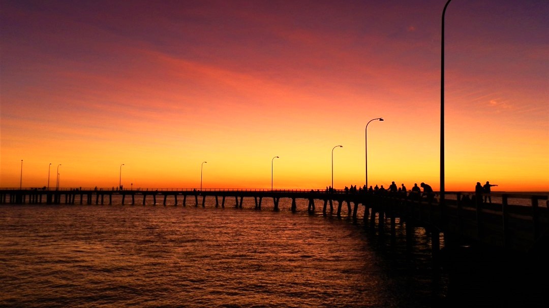 Silhouettes of people standing along Derby Jetty against a vibrant orange and purple sunset sky, creating a serene atmosphere. Watching sunsets like this is one of the popular things to do in Derby WA, offering breathtaking views and a peaceful end to the day.