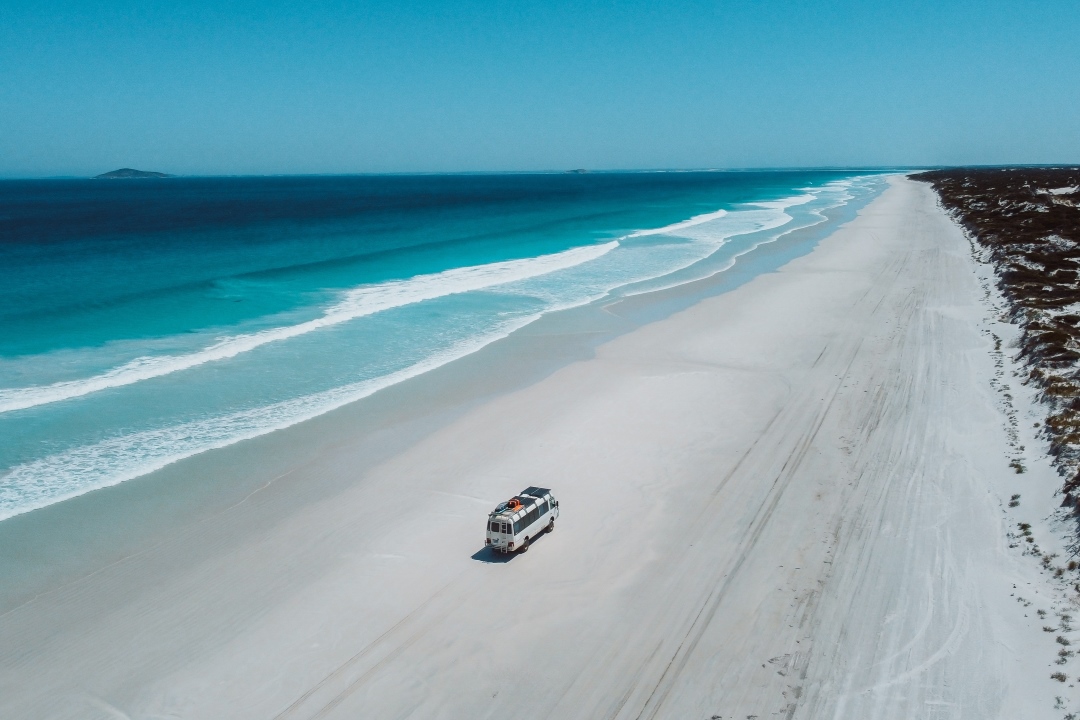 A white van driving along the vast, pristine shoreline of Cape Le Grand Beach with turquoise waves and a clear blue sky, one of the many things to do in Western Australia