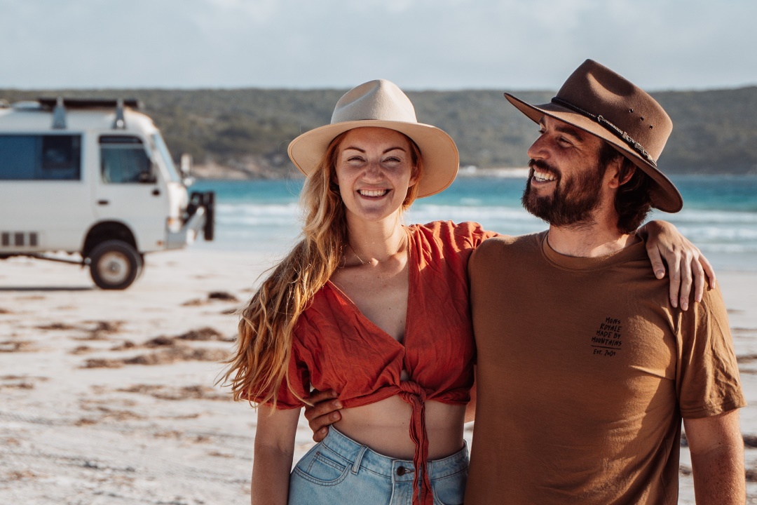 A smiling couple standing on a sandy beach with turquoise waters in the background. The woman wears a wide-brimmed hat, a red knotted top, and light denim shorts, while the man wears a brown hat and shirt. A white camper van is parked nearby, completing the scenic coastal setting.