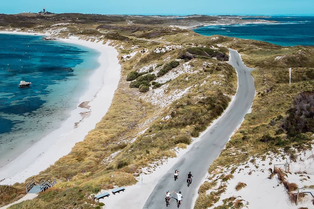 A scenic aerial view of cyclists riding along a winding coastal road on Rottnest Island, surrounded by turquoise waters, sandy beaches, and lush dunes under a clear blue sky.