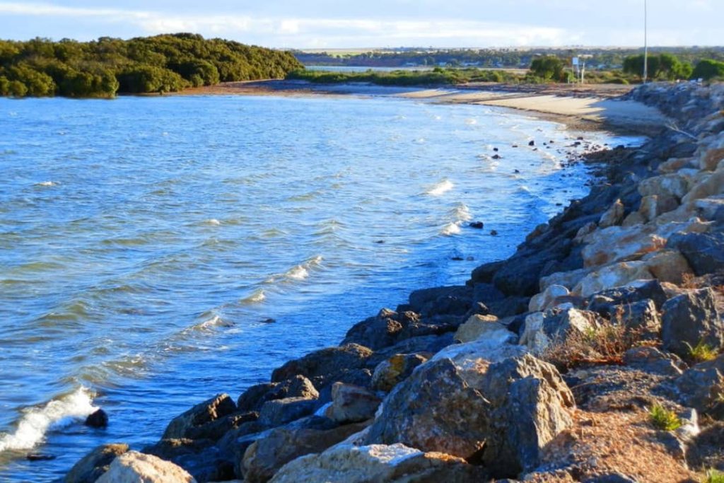 A picturesque view of Port Clinton Harbour at high tide in South Australia, with gentle waves lapping against rocky shores, mangroves in the distance, and a sandy beach curving into the background.