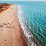 Aerial view of Gantheaume Point, Broome, showing a long stretch of golden sandy beach meeting turquoise ocean waters. The end destination for a road trip from Darwin to Broome