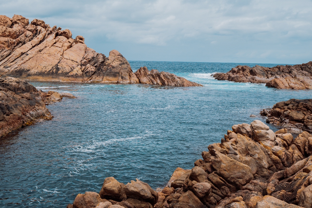 A view of rugged rock formations rising from the turquoise ocean at Canal Rocks, with waves gently breaking against the rocks under a cloudy sky