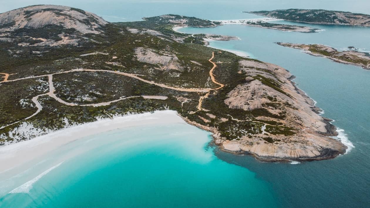 Aerial view of Wharton Beach in Esperance, Western Australia, showcasing clear turquoise water, white sandy shores, and rugged coastal cliffs surrounded by green vegetation.