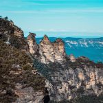 Scenic view of the Three Sisters rock formation in the Blue Mountains, Australia, surrounded by lush greenery under a bright blue sky. The best time to visit this iconic location is spring, when the flowers bloom and the weather is mild.