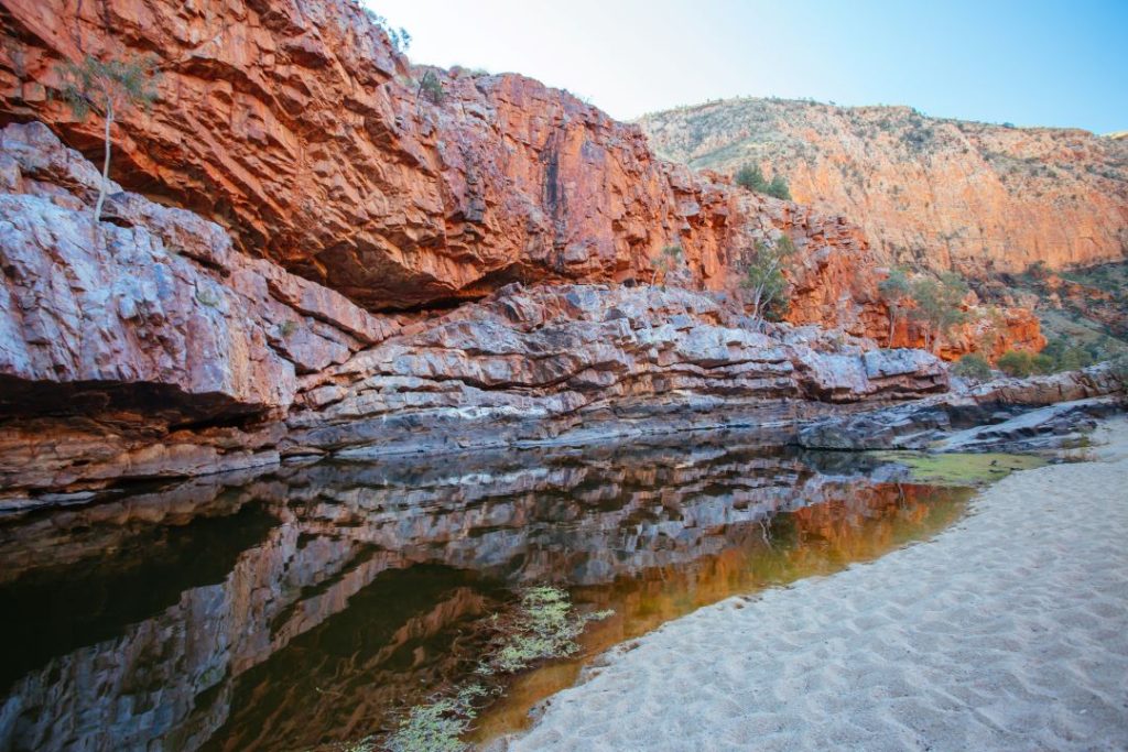 The red rock walls of Ormiston Gorge in the Northern Territory, Australia, reflected in a calm pool of water surrounded by sandy banks.