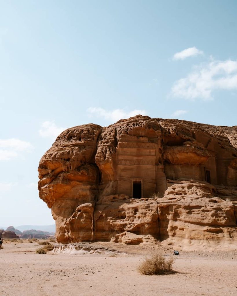 A detailed view of an ancient carved rock tomb in Hegra, AlUla, Saudi Arabia, showcasing intricate patterns and architectural features carved into the sandstone. The desert setting with scattered shrubs enhances the historical atmosphere under a clear blue sky.