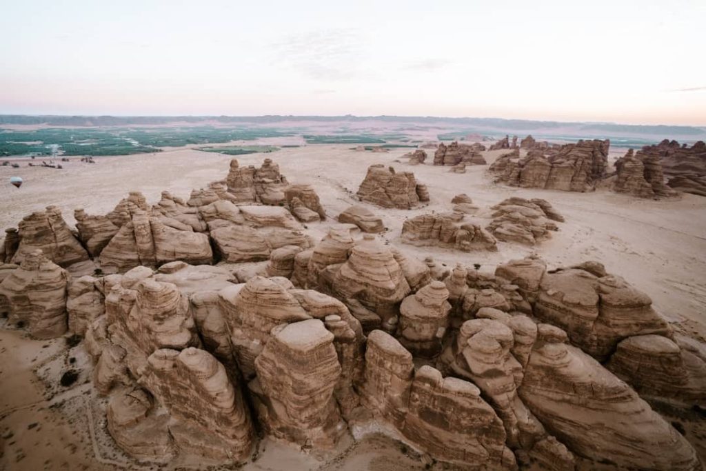 An aerial view of the sandstone rock formations in Hegra, AlUla, Saudi Arabia, showcasing the intricate natural structures surrounded by sandy desert terrain. In the distance, green agricultural fields contrast with the arid landscape under a soft pastel sky.